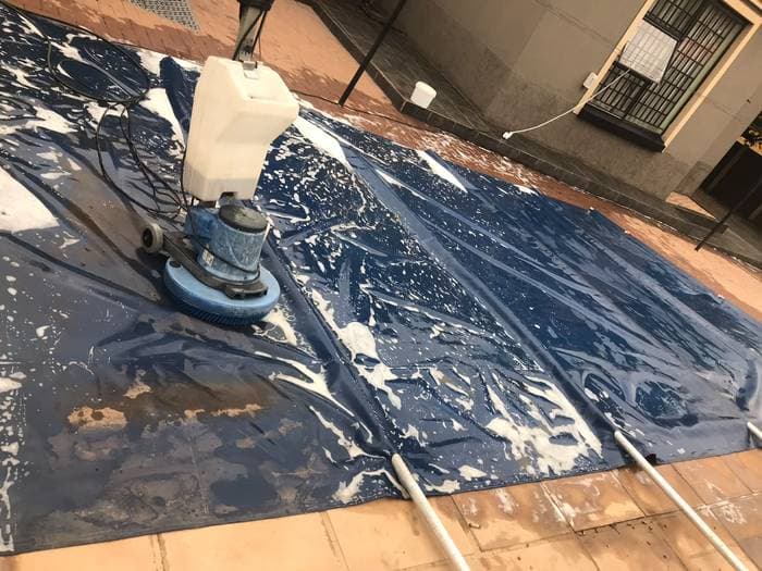 A rotary cleaning machine scrubbing a large soapy pool cover on site.
